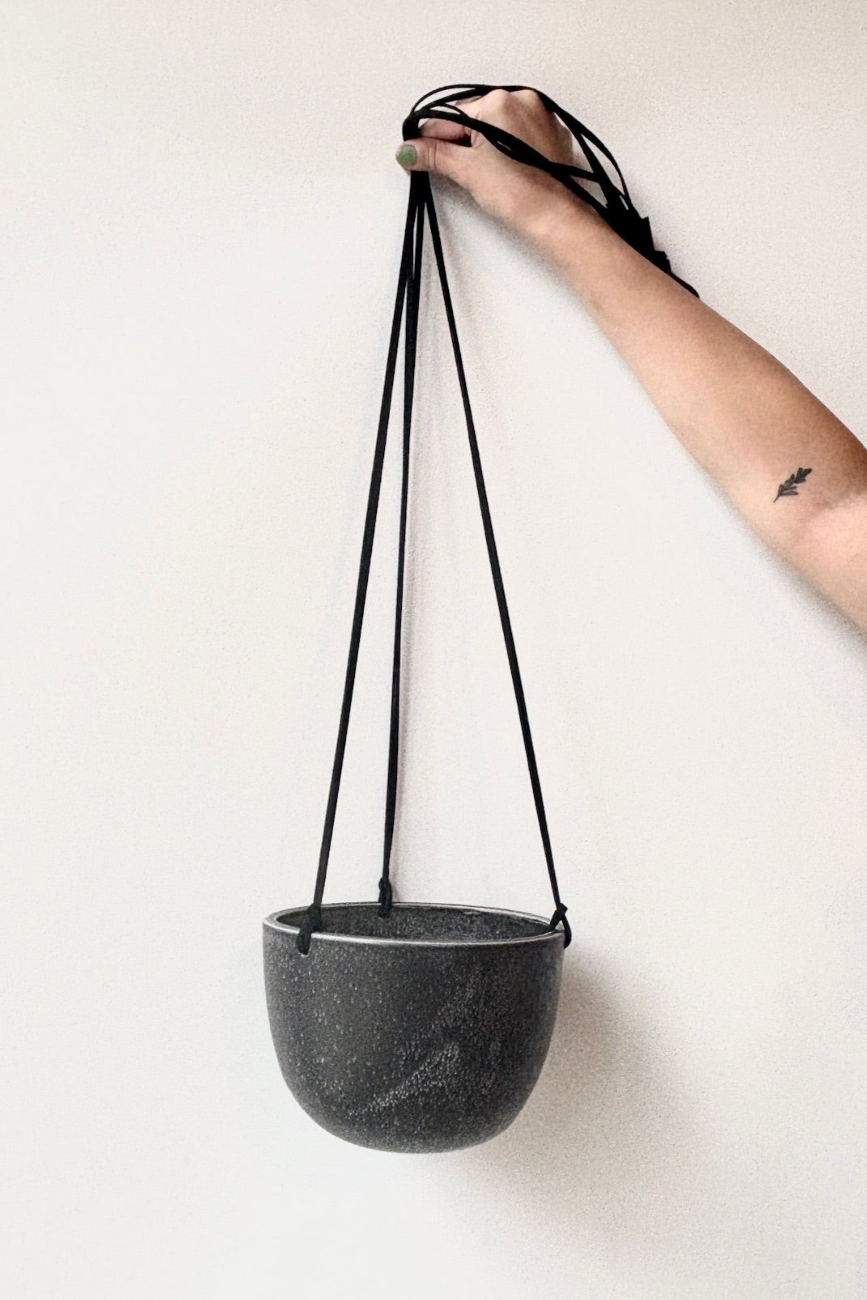 Black speckled ceramic hanging planter with a black strap held by a hand on a white background ©Sprout Home