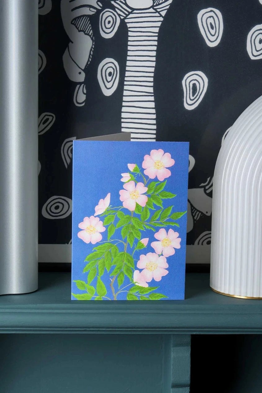 Dog rose greeting card by Stengun Drawings  standing on a shelf with a black and white background. 