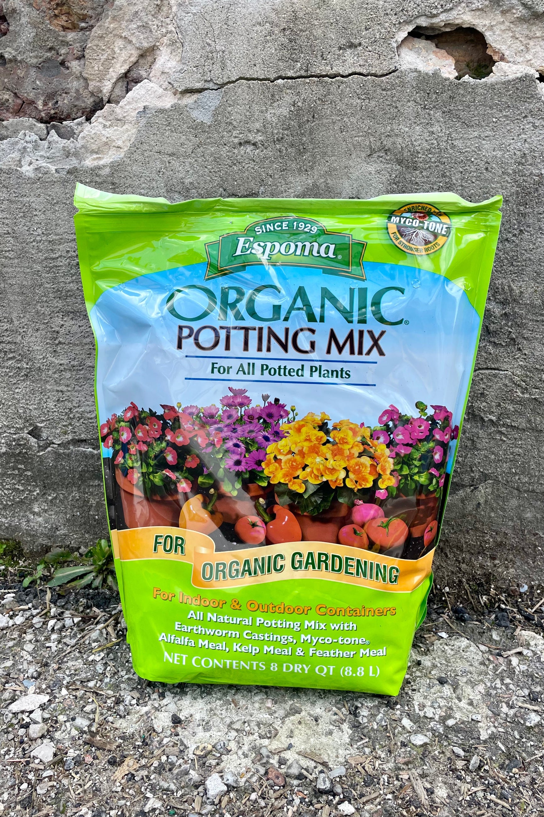 Photo of a plastic bag of organic potting mix against a cement wall. The bag is bright green with a middle band of illustration and text showing the product name and images of flowers and vegetables with the text "for organic gardening." The bottom text shows the contents and volume of the bag.