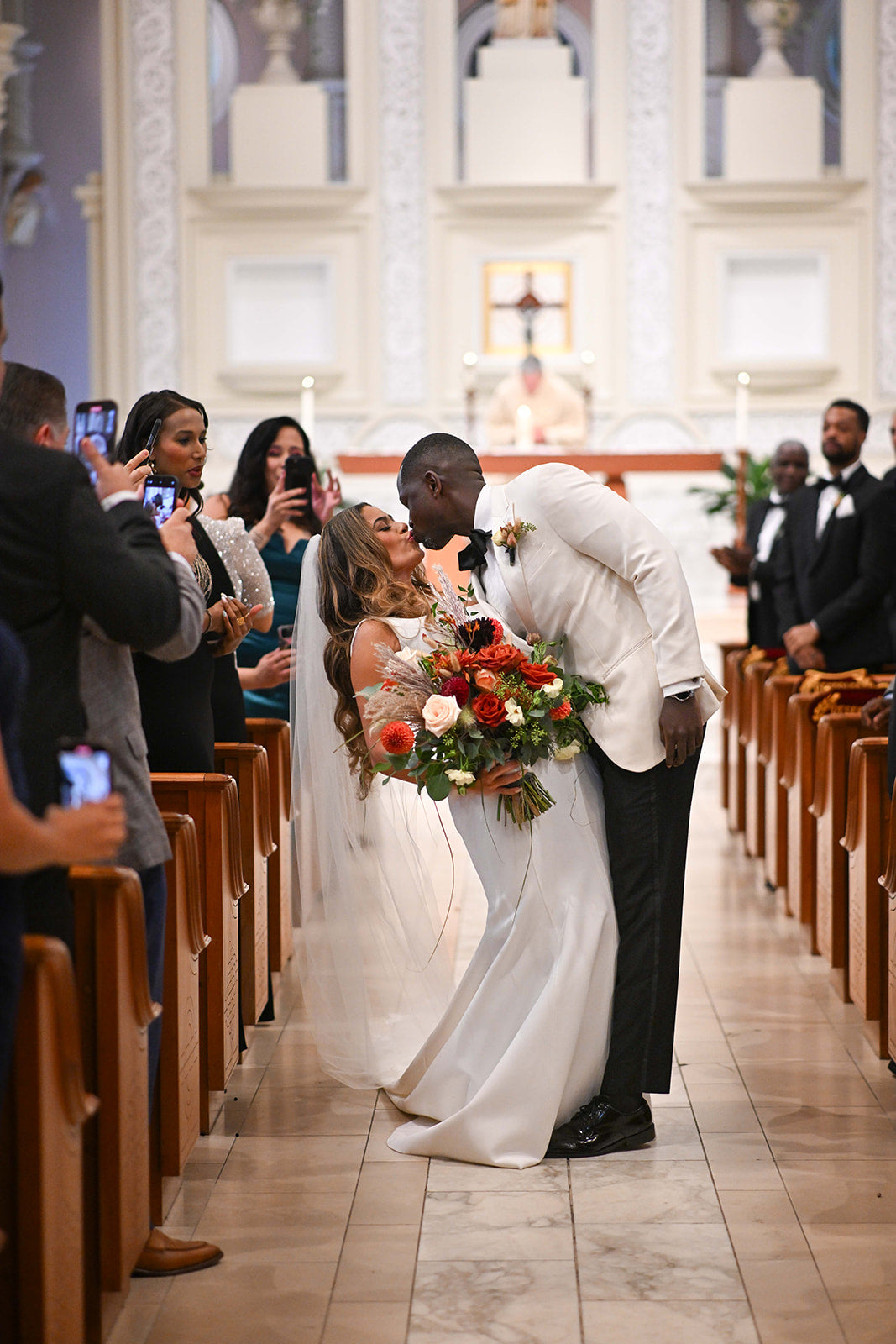 Sam & Kwaku's wedding. Bride holding a large earthy colored bouquet with huge flower pops standing and kissing in the aisle of a church.  Floral by Sprout-home. Photograph by Mack Julion