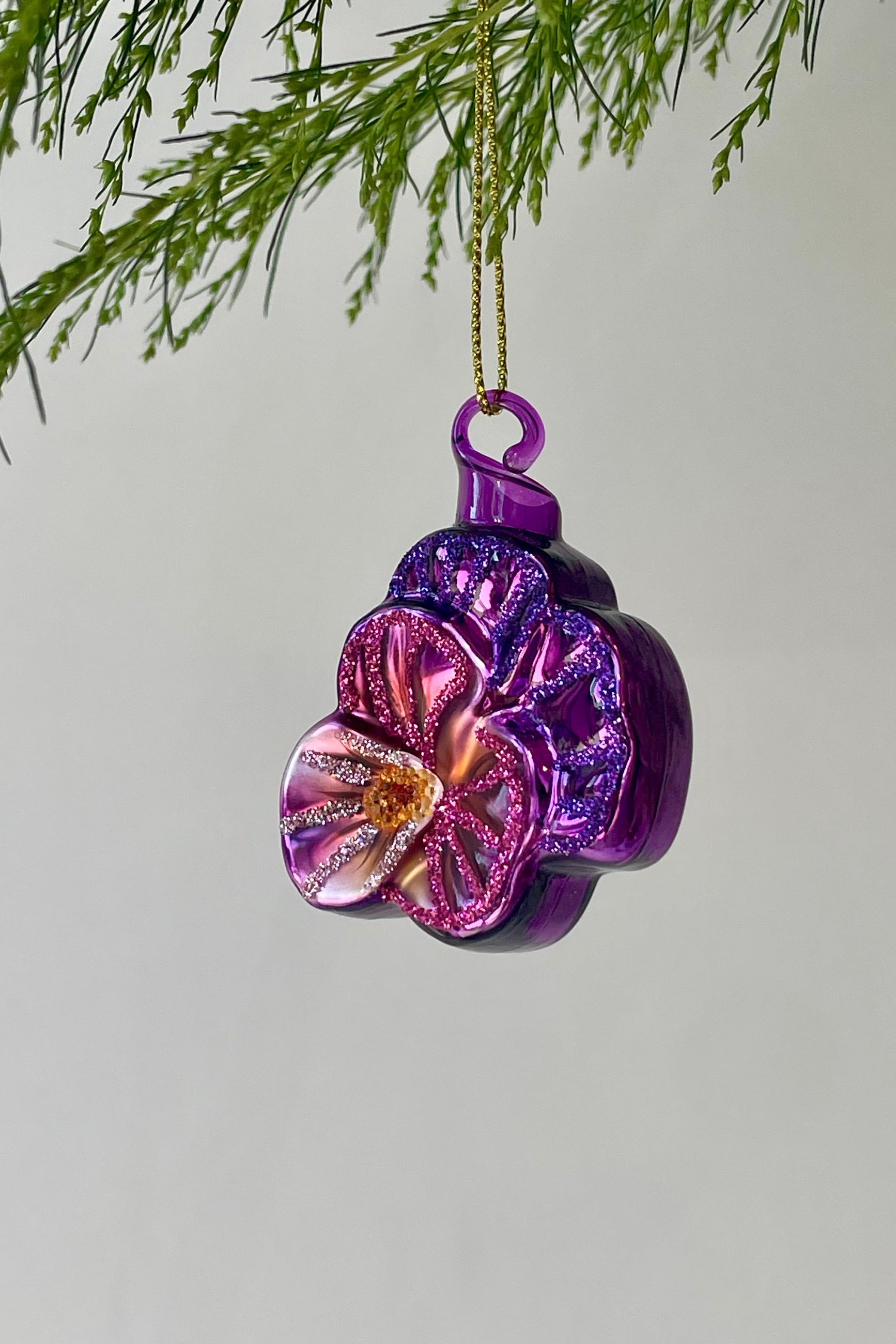 A purple floral ornament shaped like a violet with glitter hangs from a branch in front of a gray wall ©Sprout Home