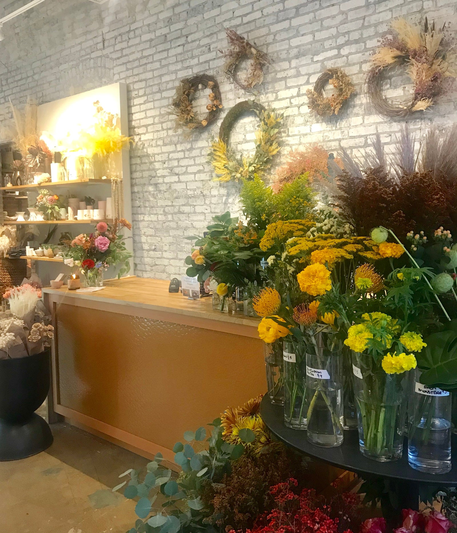 Sprout-home Floral store counter with dried floral wreaths on the white brick wall and fresh floral in glass vases in the right corner