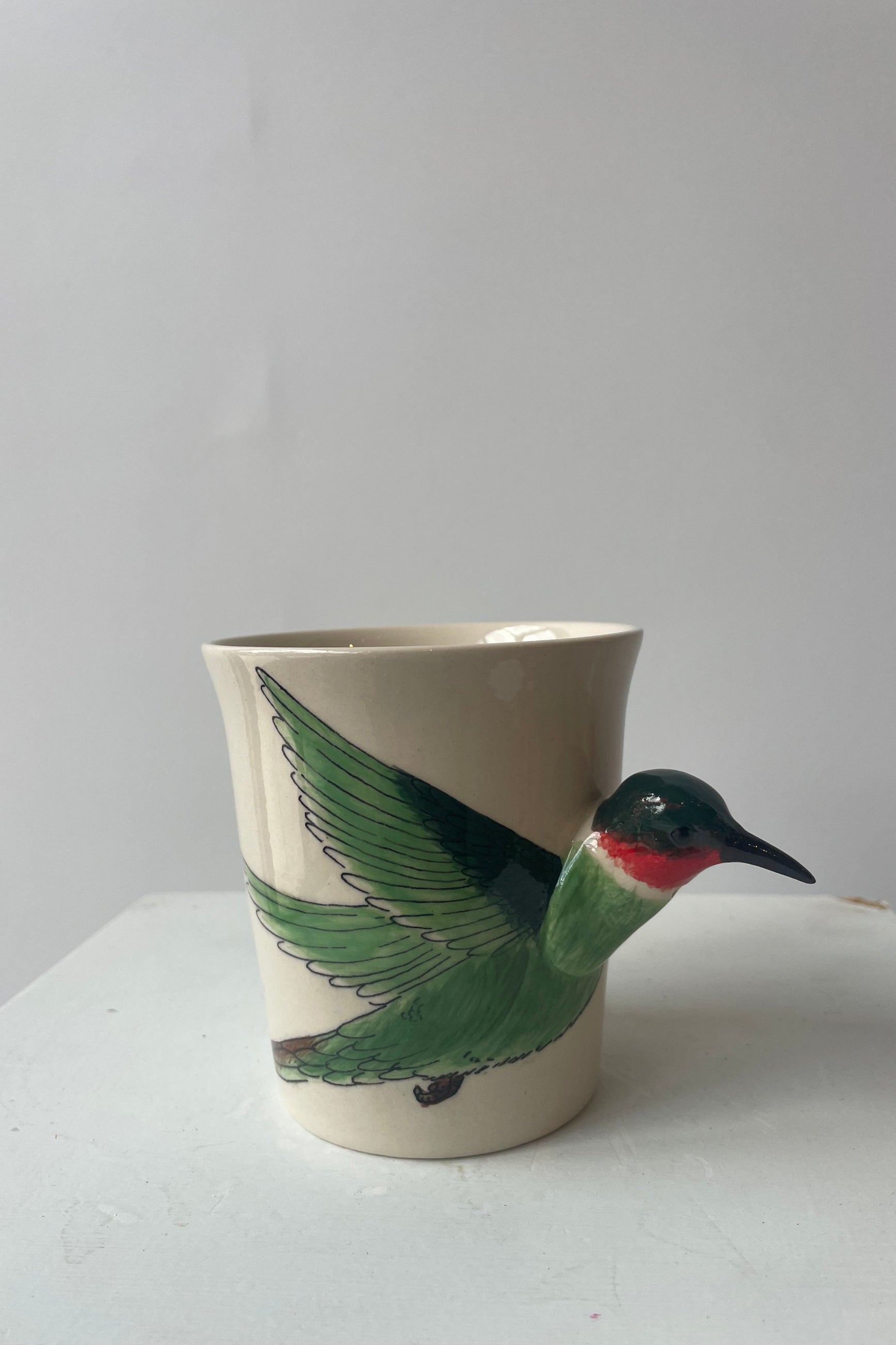 A white mug sits on a white table in a white room at Sprout-home. A green and ruby throated hummingbird shapes the side and handle of the mug. The mug sits upright.