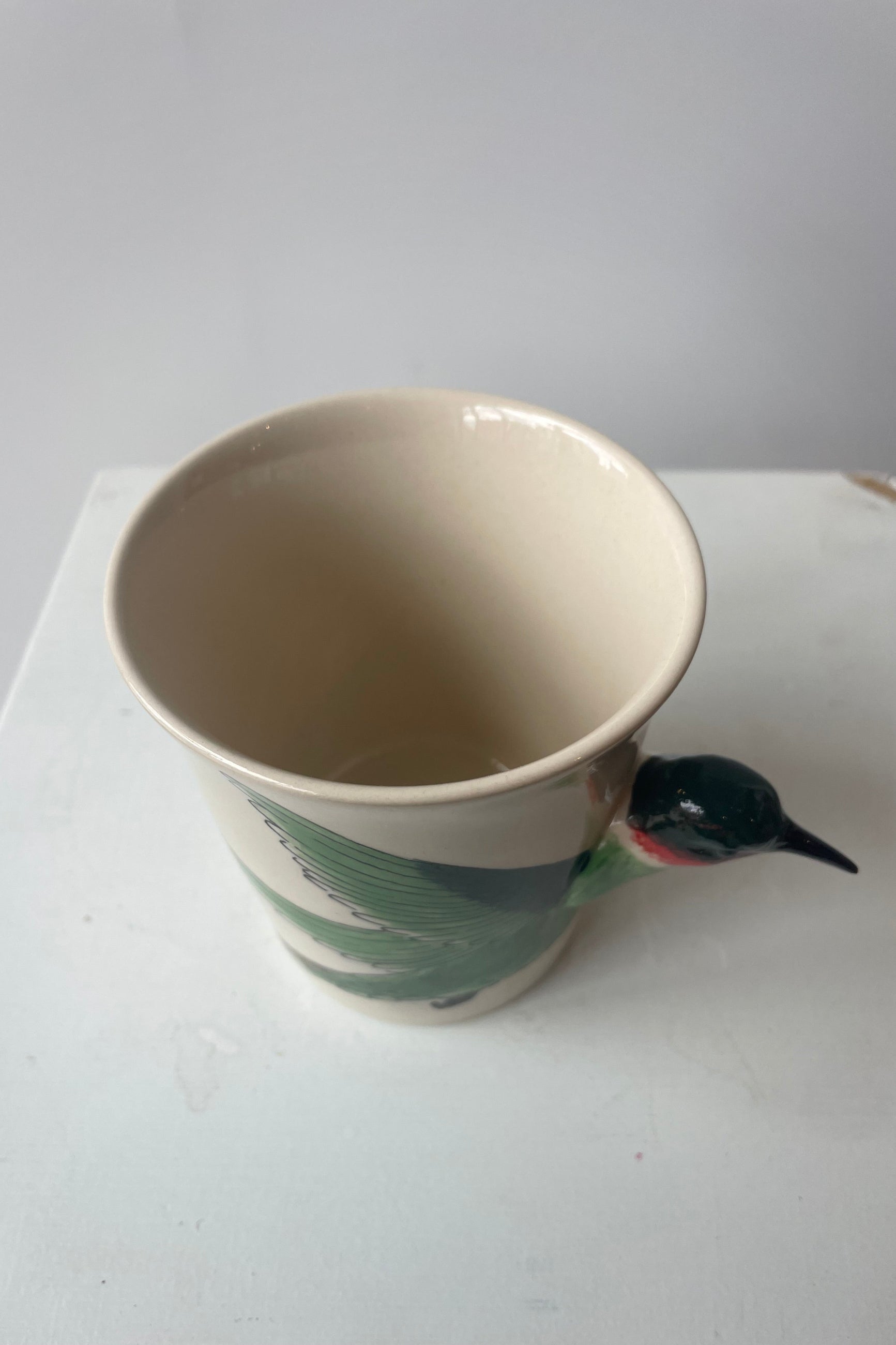 A white mug sits on a white table in a white room at Sprout-home. A green and ruby throated hummingbird shapes the side and handle of the mug.  The view is looking down into the mug.