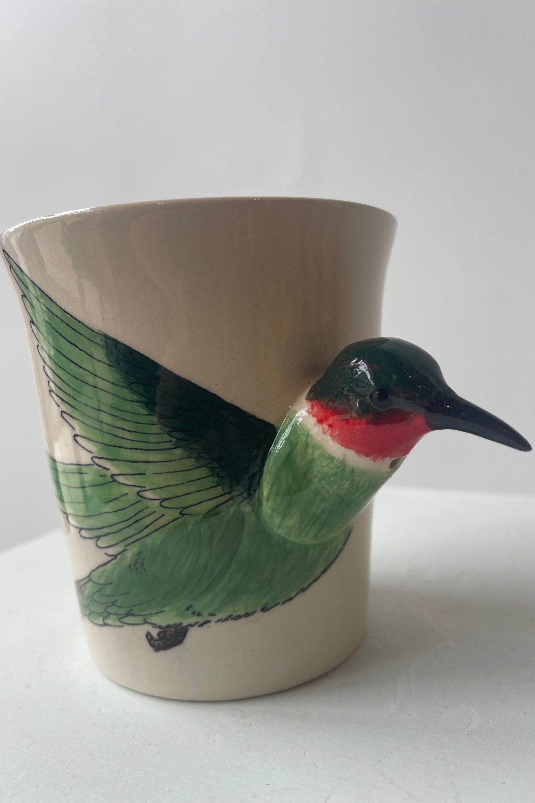 Close view of a white mug sits on a white table in a white room at Sprout-home. A green and ruby throated hummingbird shapes the side and handle of the mug. 