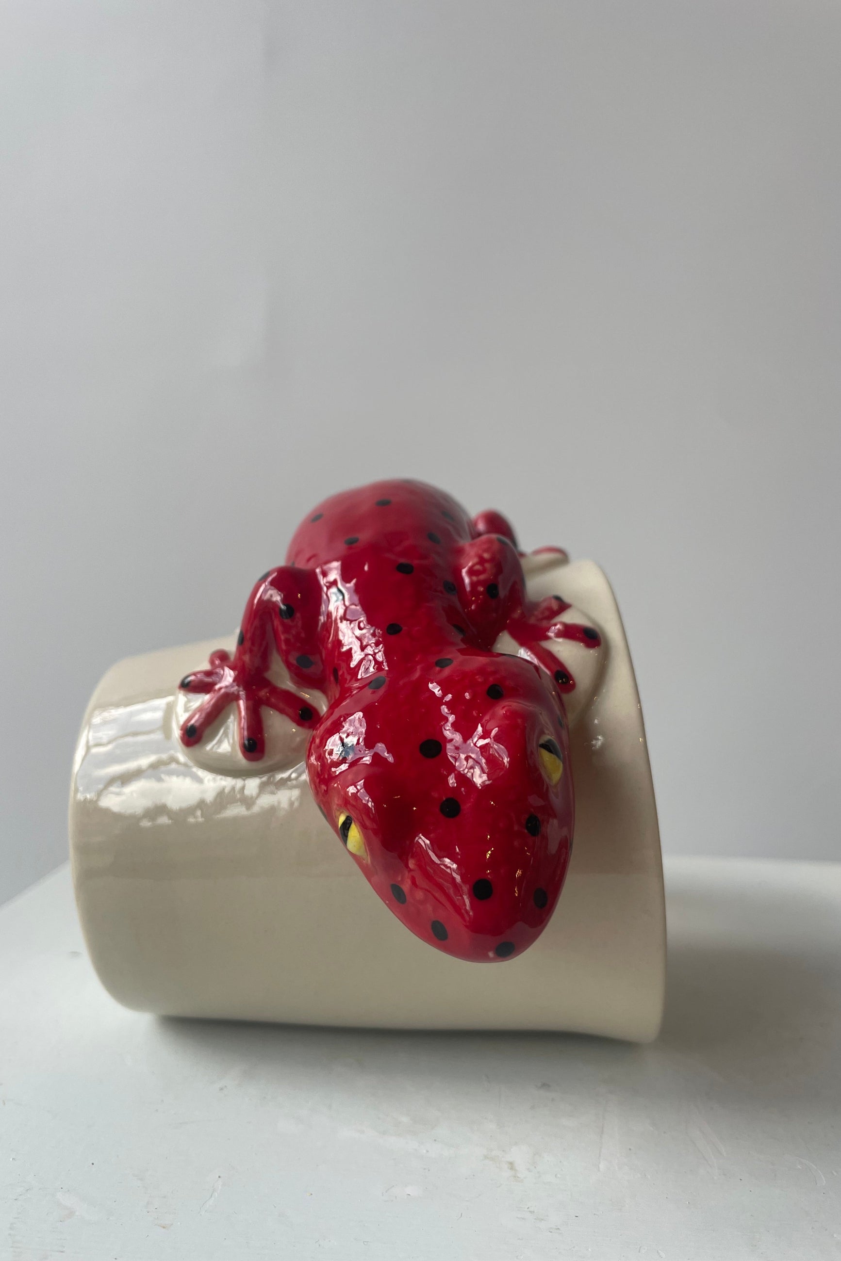 Photo of a white mug on a white table in a white room. A red salamander shapes the body and handle of the mug. The mug sits on its side at Sprout-home.