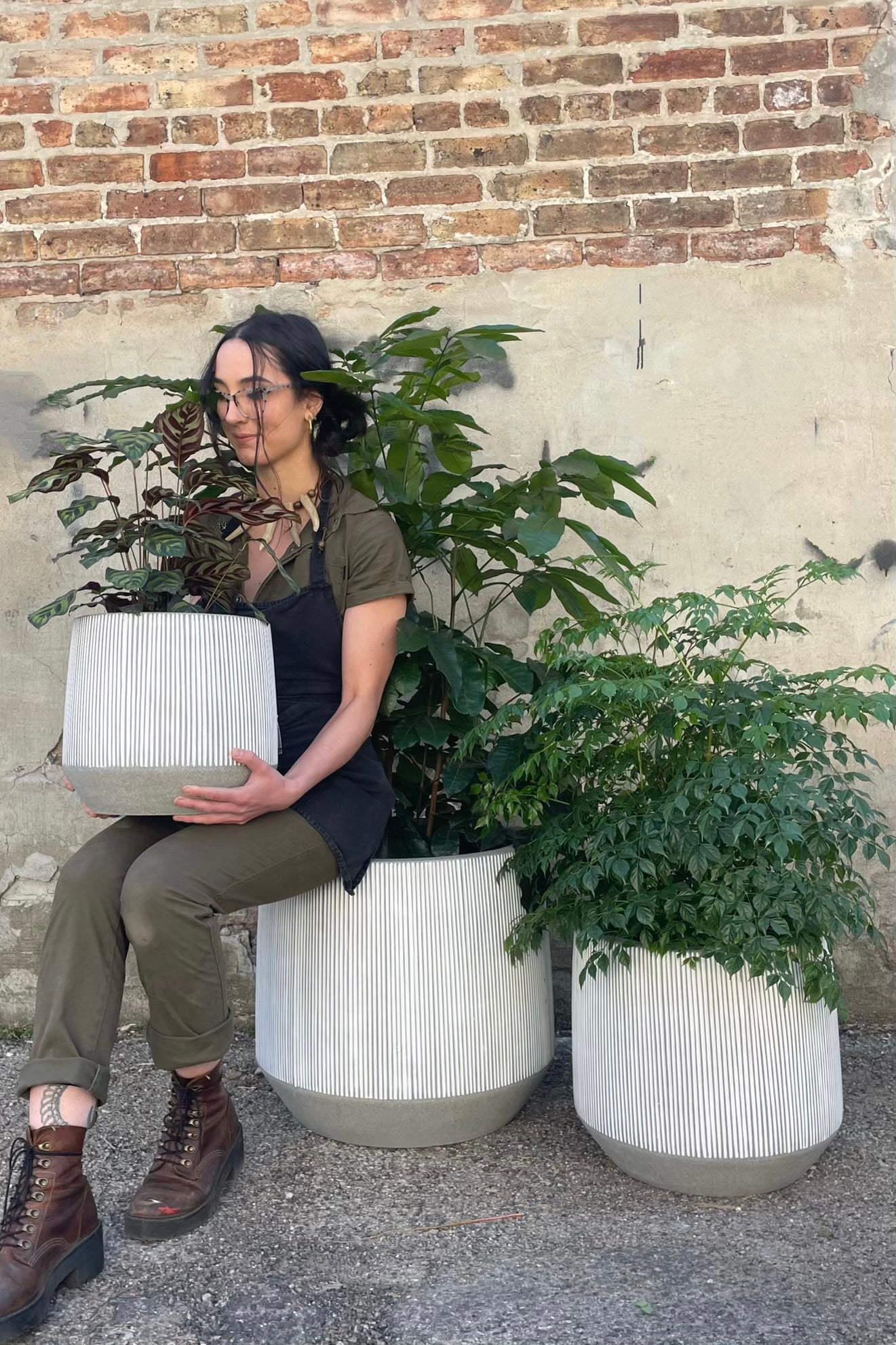 A person sitting with three sizes of white and gray striped  Harith pots and plants against a cement and brick weall.