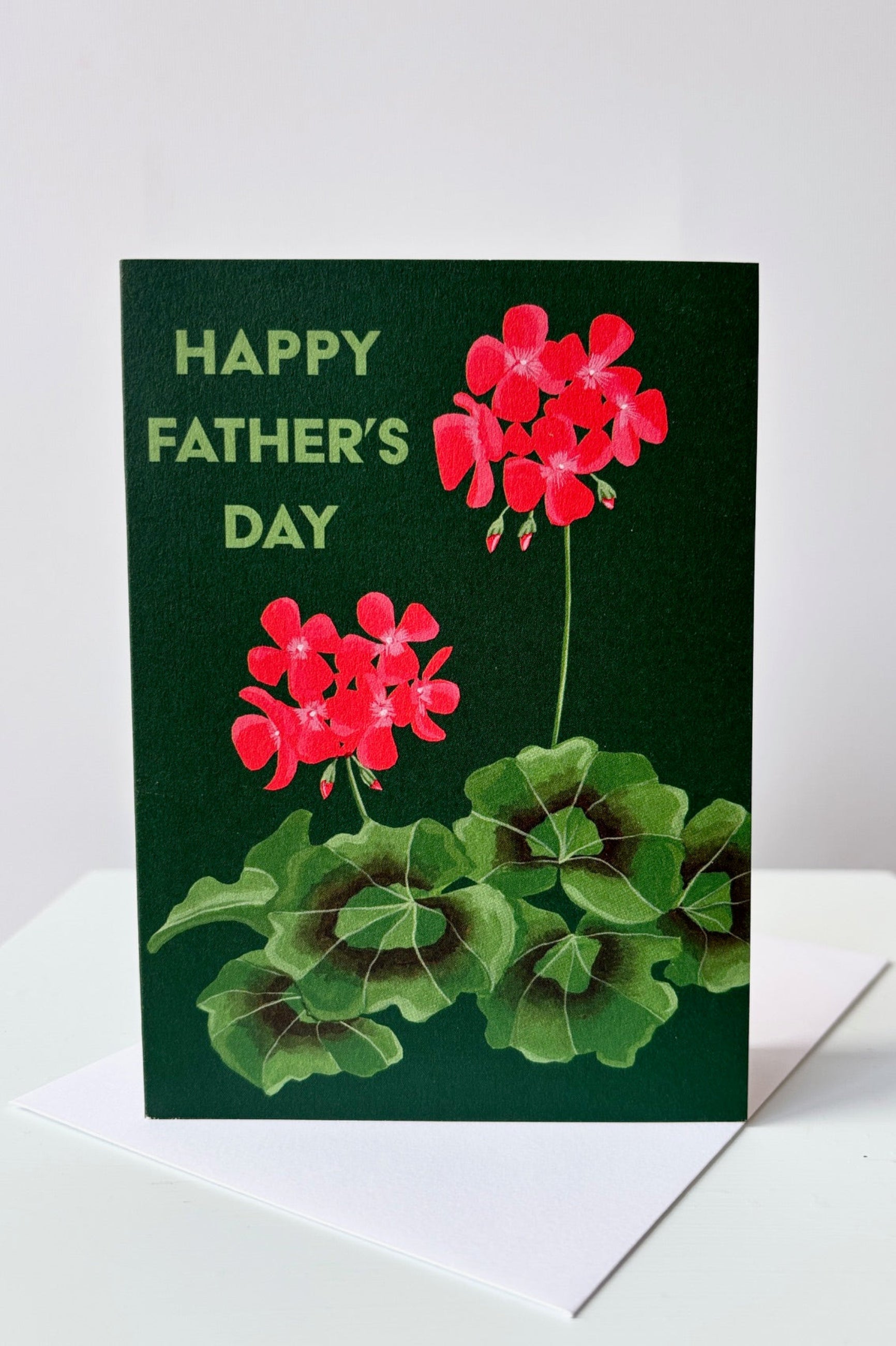 Greeting card with image of red flowering zonal begonia with text that says Happy Father's Day on a green background. Shown with envelope ©Sprout Home