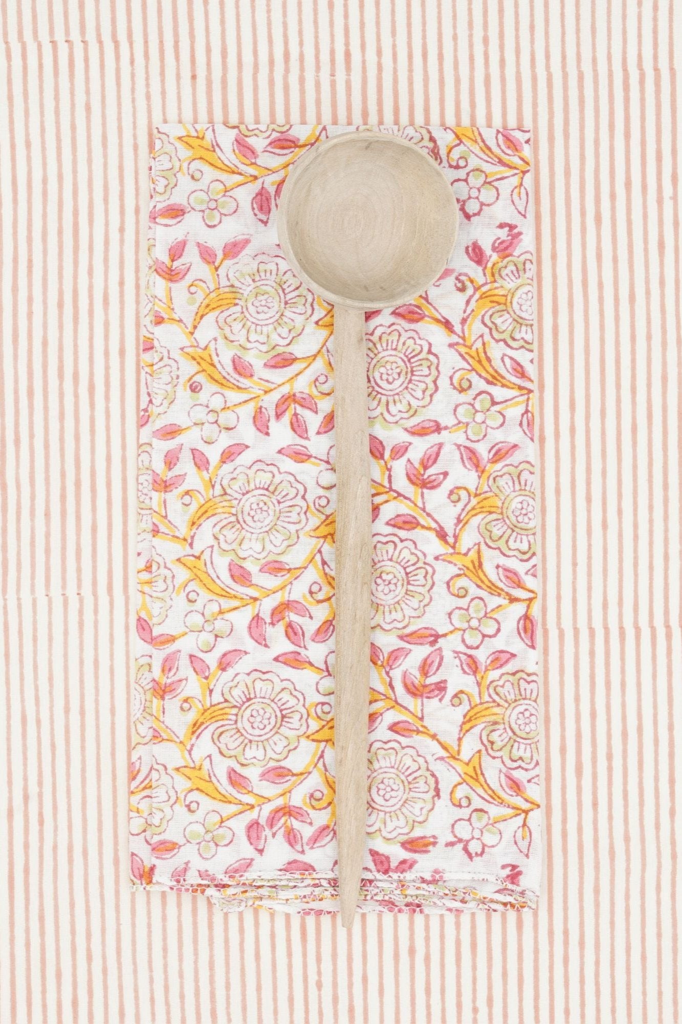 Suzette Frangipanier Jaipur napkin folded in to a rectangle, wood spoon on top and on a striped background. 