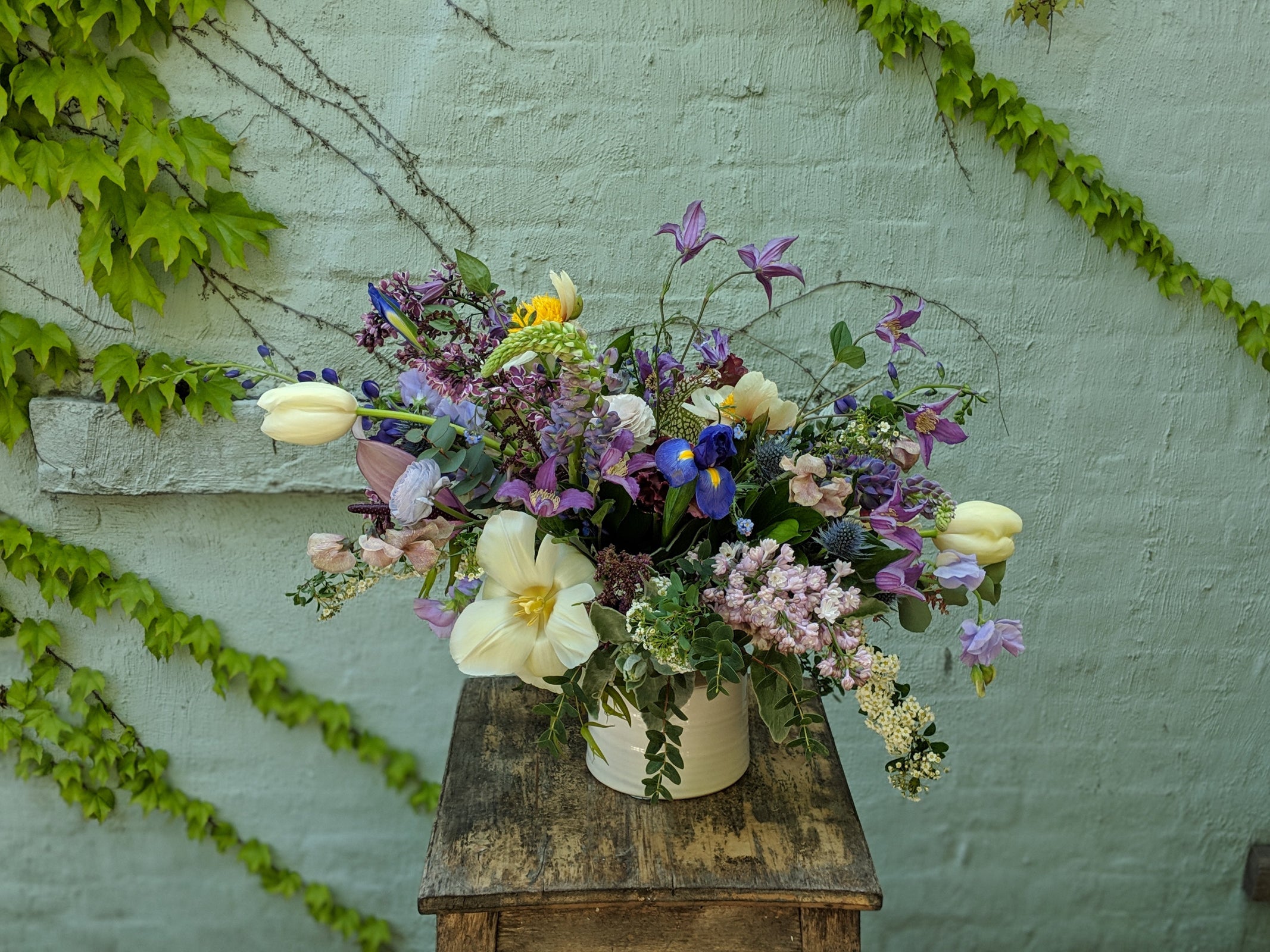Floral arrangement by Sprout-home focusing in purples and blues against a blue green wall with ivy. 